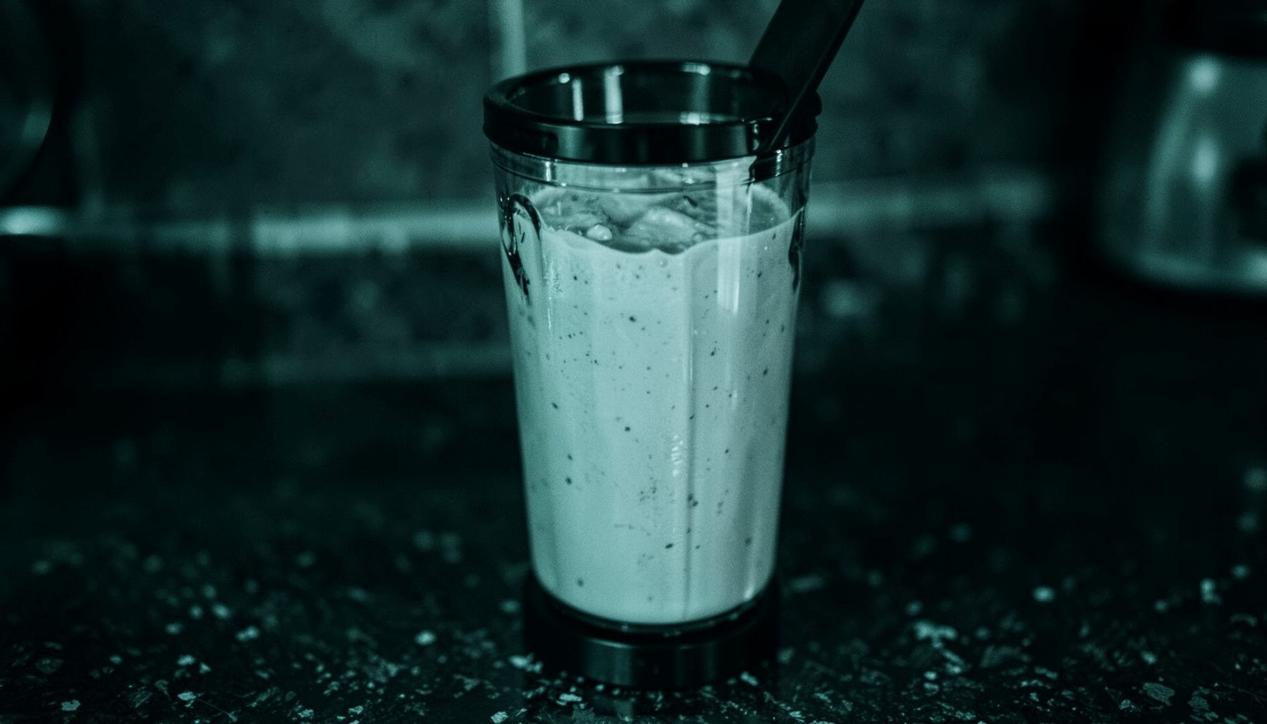 Duotone photograph of a blender bottle with creamy shake liquid against a dark kitchen counter, deep teal and black tones