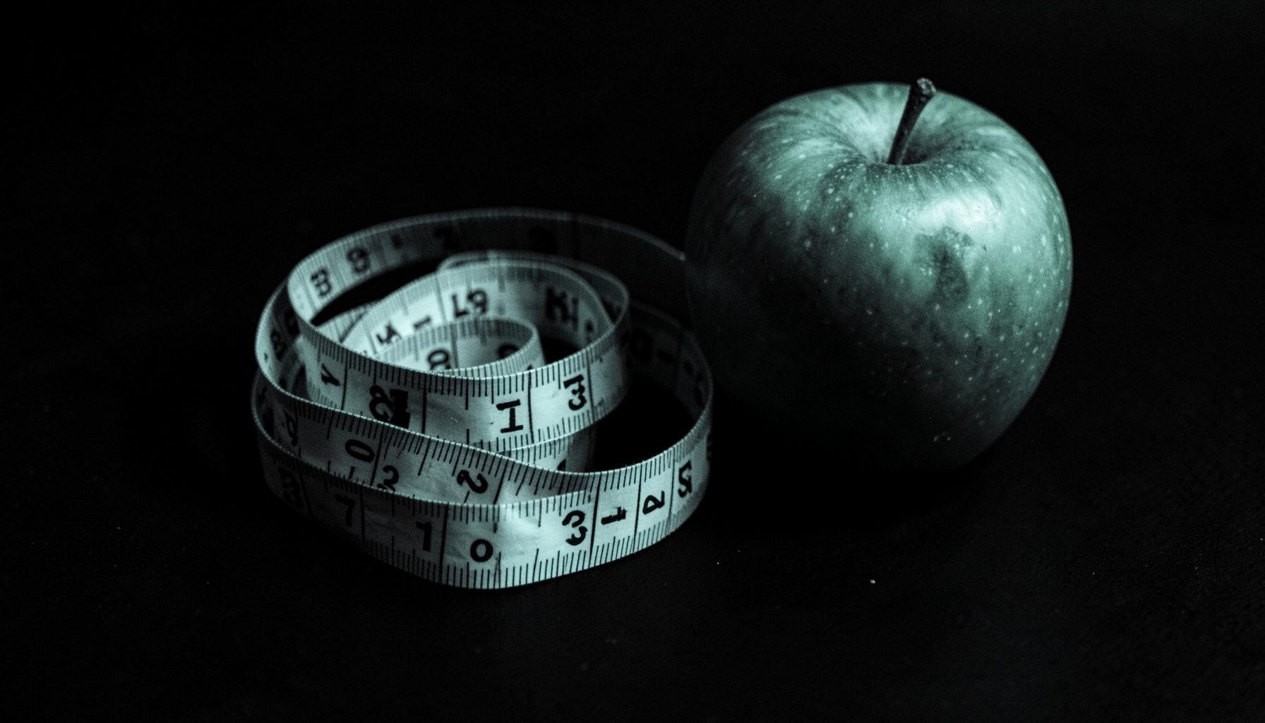 Dark duotone photograph of a measuring tape loosely coiled beside a green apple on a matte black surface, moody and focused