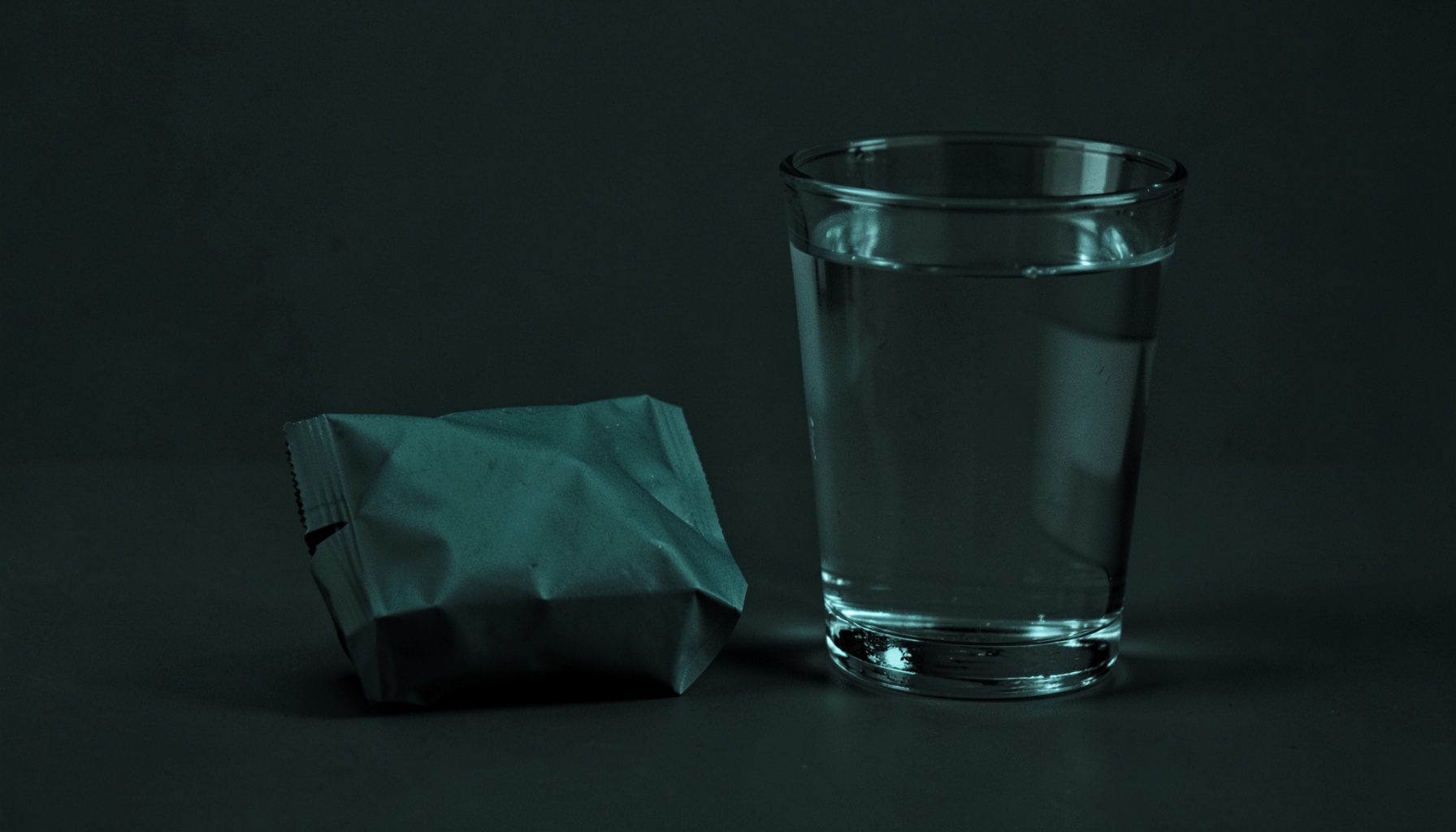 Duotone photograph of a single-serving shake packet next to a glass of water, minimalist dark composition