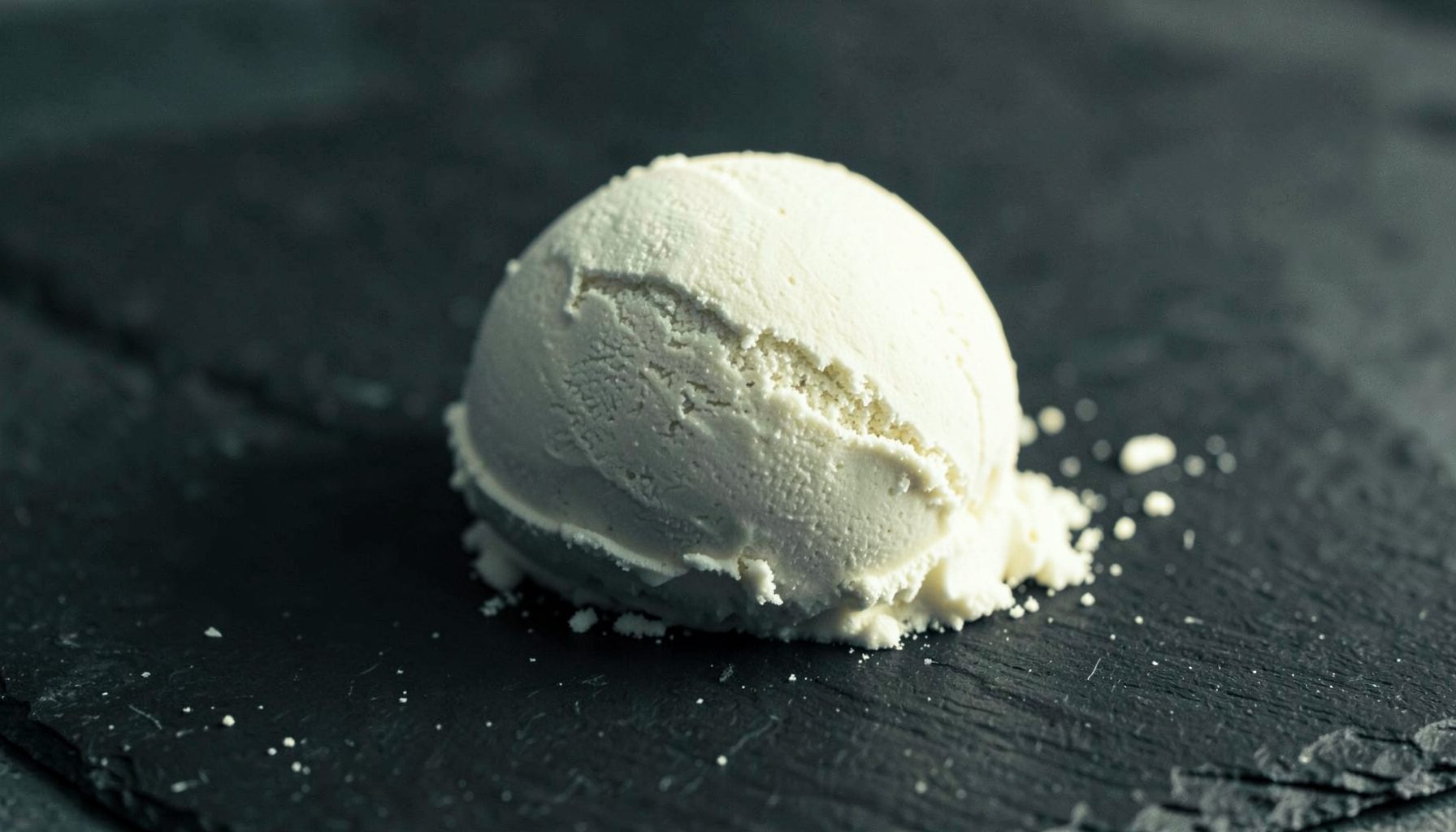 Duotone photograph of a scoop of vanilla protein powder resting on a dark slate surface, warm cream and charcoal tones
