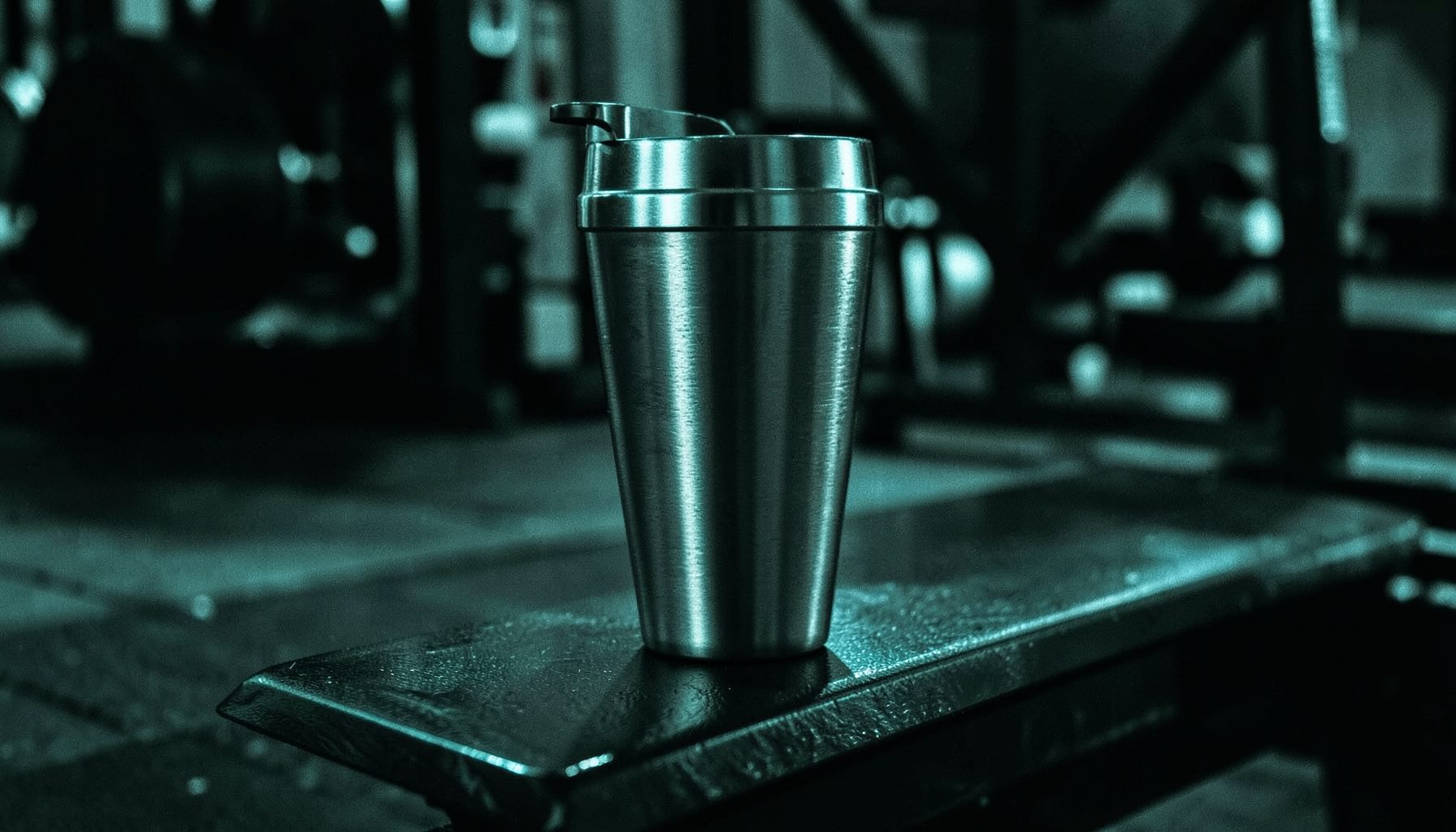 Duotone photograph of a stainless steel shaker cup on a gym bench, dark moody lighting with teal accents