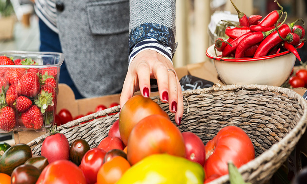 Close-up of a hand with red nail polish reaching into a basket of fresh tomatoes, with strawberries and chili peppers nearby