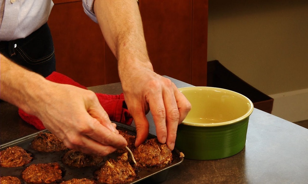 Hands placing food patties into a baking tray next to a green bowl on a kitchen counter, warm brown tones, practical cooking mood