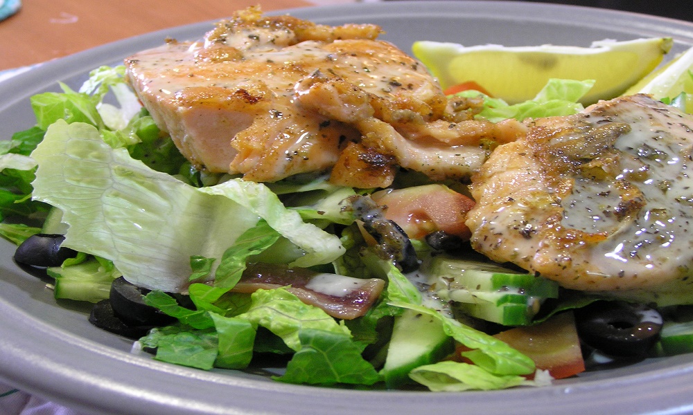 Plate of grilled meat and fish over a green salad with cucumbers, tomatoes, and olives, appetizing warm tones